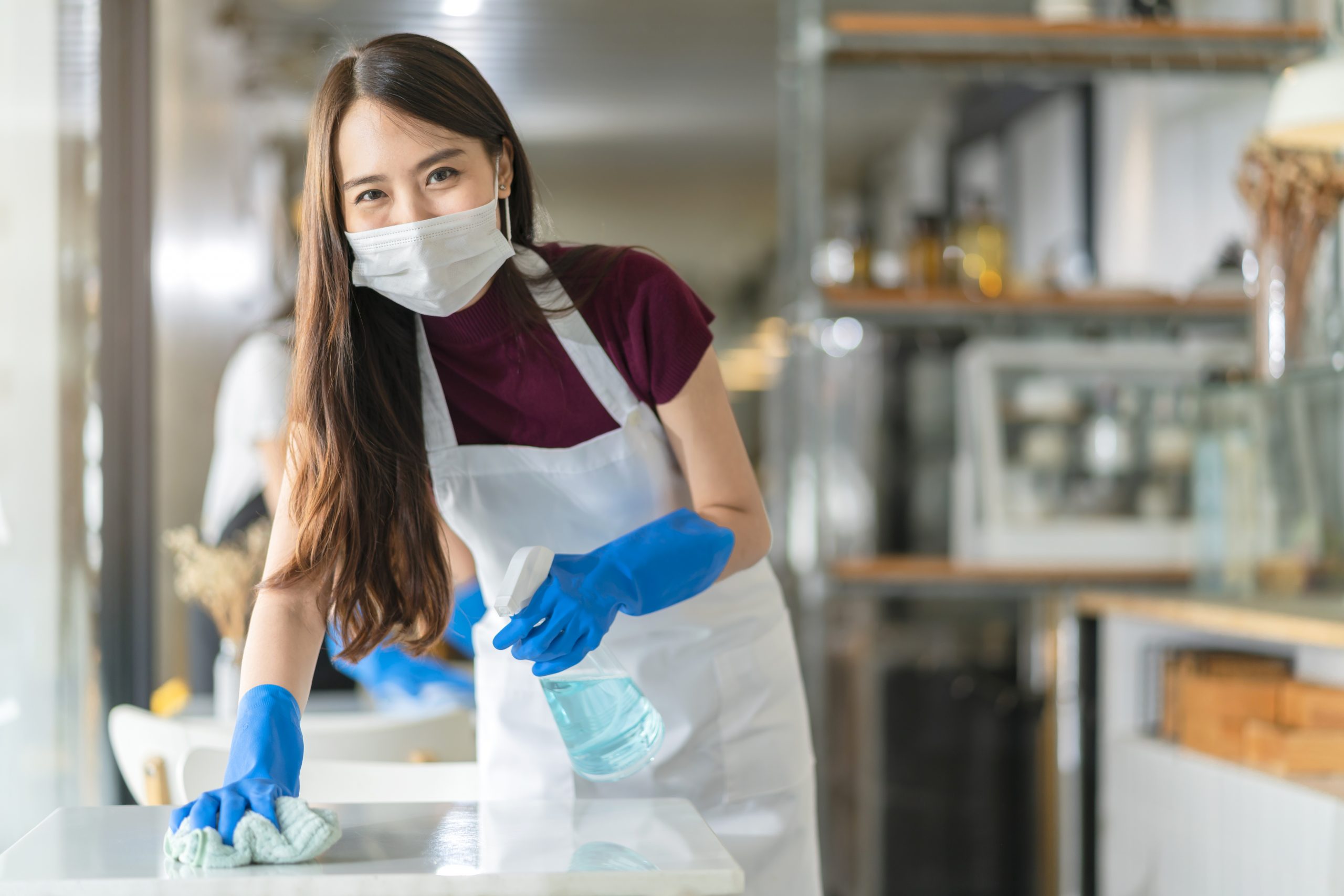 Asian staff waitress wearing protection face mask apron rubber glove cleaning table with disinfectant alcohol spray prevent infection coronavirus cafe coffee shop new normal concept